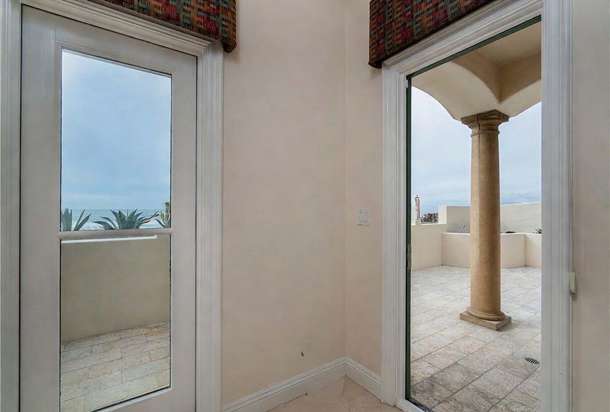 This interior view captures a transition space featuring two glass-paned doors that open onto a tiled outdoor terrace. The neutral-toned walls and light-colored flooring create a bright, airy atmosphere, while the open door on the right reveals a classic architectural column on the balcony. The perspective highlights the seamless connection between the indoor living area and the outdoor space, emphasizing the coastal view visible through the glass.