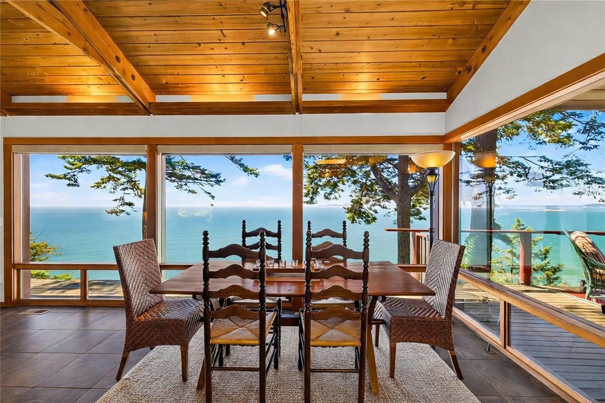 This dining room features a rustic wooden table surrounded by four woven chairs, set upon a textured area rug. The space is defined by expansive, floor-to-ceiling windows that provide a panoramic view of the ocean and surrounding trees. A warm, wood-paneled vaulted ceiling with exposed beams and track lighting adds a cozy, cabin-like atmosphere to the bright, airy room.