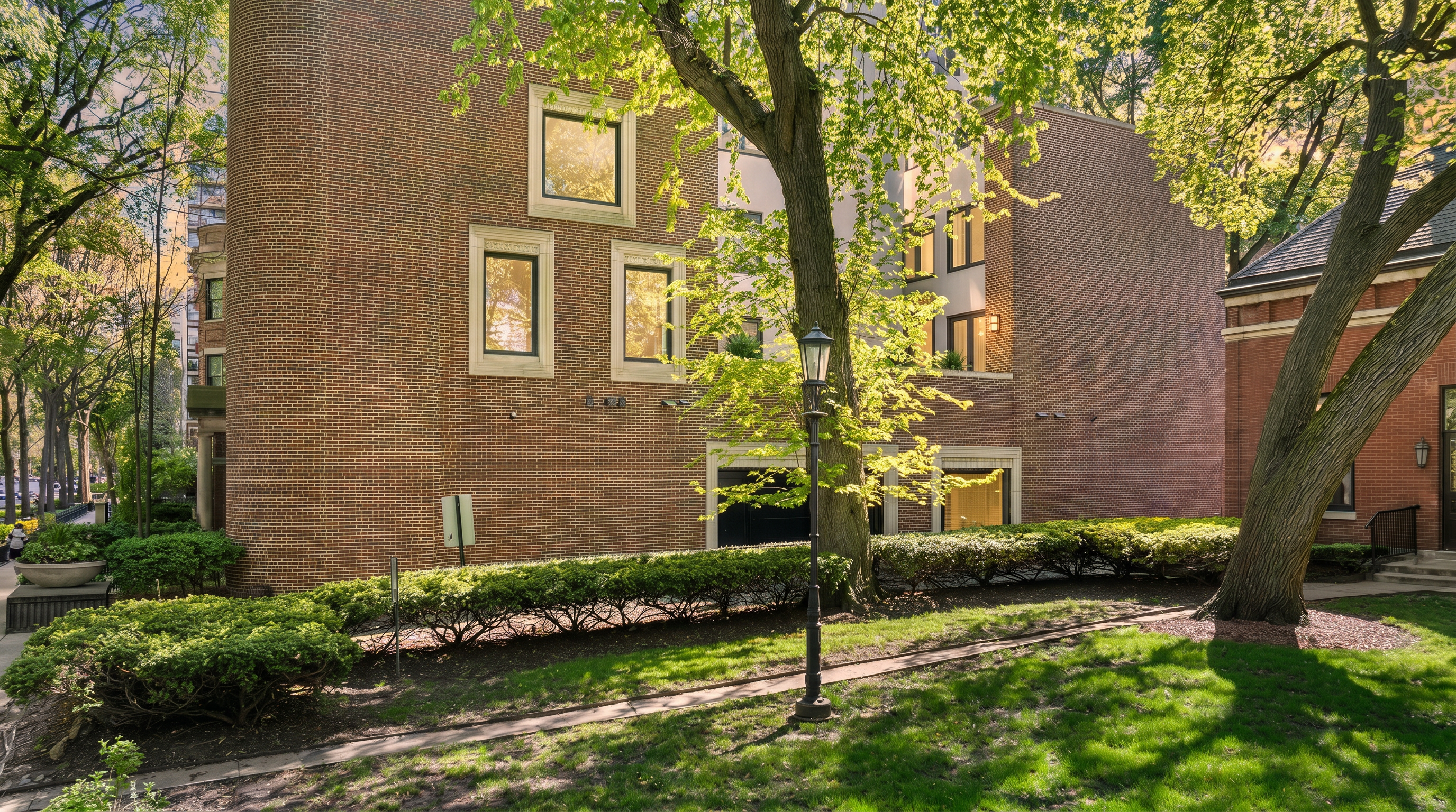 This side view of a multi-story brick building showcases a distinctive curved corner design and a series of rectangular windows with light-colored stone trim. A mature tree stands prominently in the foreground, casting dappled shadows across a manicured lawn and a neatly trimmed hedge that runs along the base of the structure. The scene is captured from a ground-level perspective, emphasizing the lush landscaping and the architectural contrast between the brick facade and the surrounding greenery.