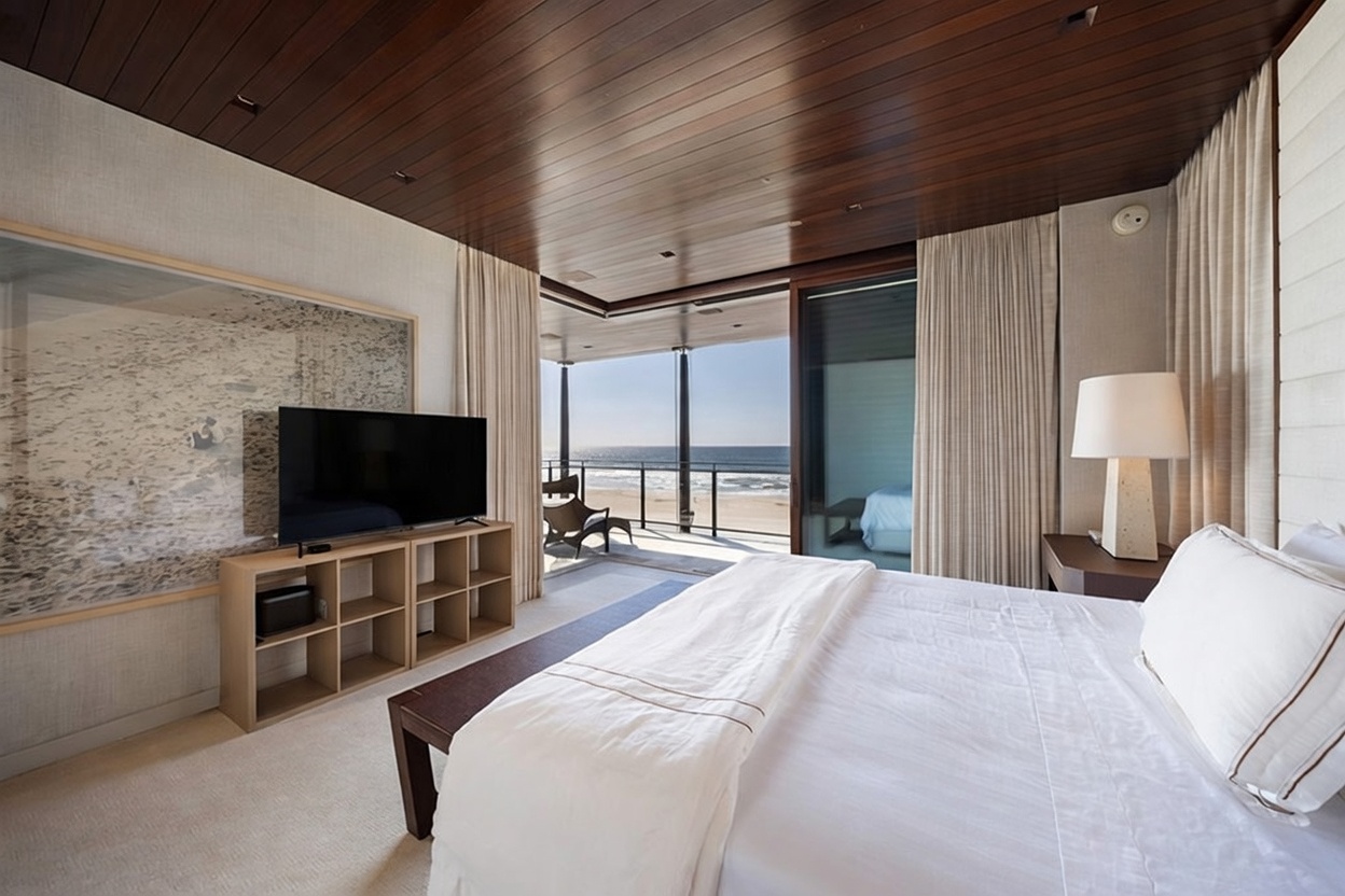 This primary bedroom features a sophisticated, modern design with a warm wood-paneled ceiling and neutral-toned walls. A large bed sits in the foreground, while a media console with a television is positioned against a large, textured wall art piece. The room opens directly onto a private balcony through expansive glass sliding doors, offering an unobstructed, panoramic view of the ocean and beach.