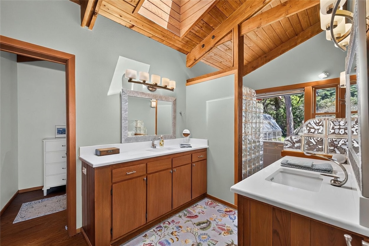 This primary bathroom features a dual-vanity layout with light wood cabinetry and white countertops, set against soft green walls. The space is highlighted by a vaulted wood-paneled ceiling with a skylight and a glass block partition that separates the vanity area from the shower. A floral-patterned rug adds a touch of color to the floor, while large windows provide a view of the surrounding greenery.
