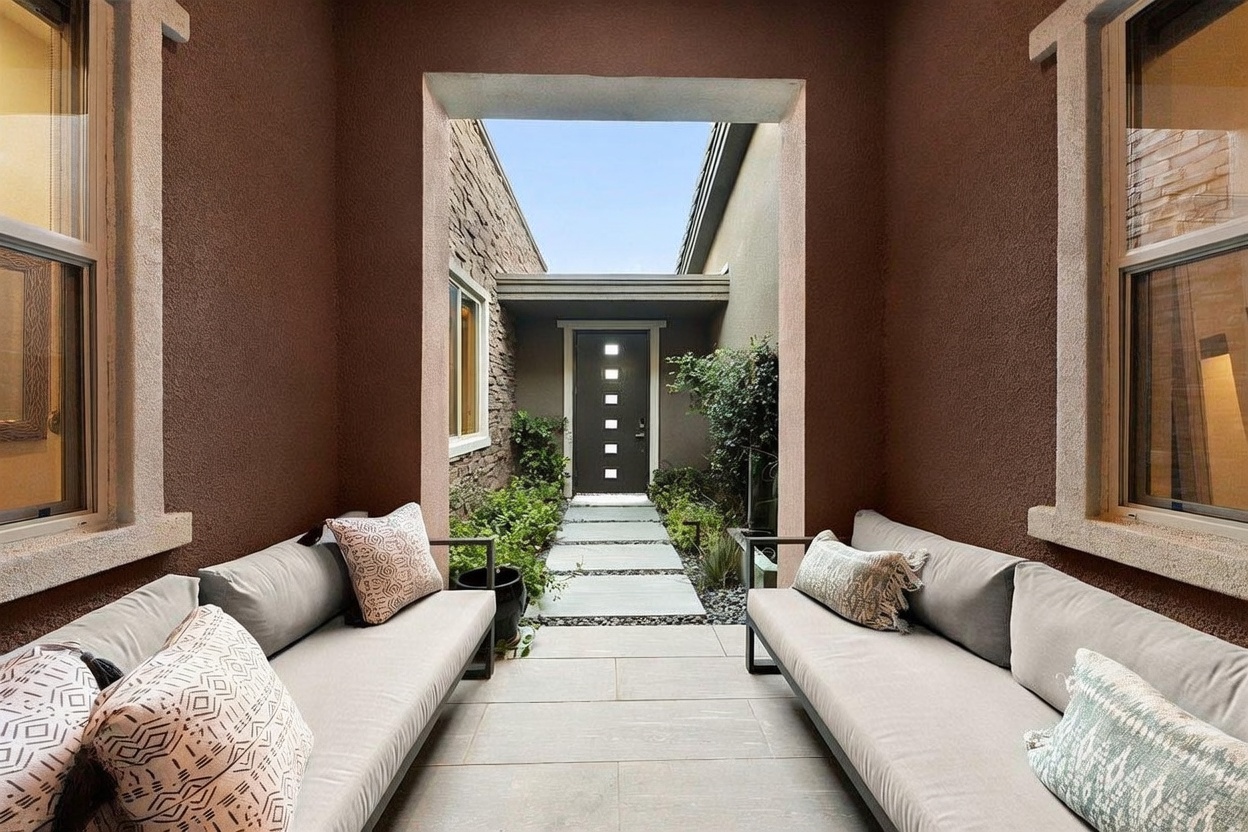 This inviting courtyard entryway features a symmetrical layout with two long, cushioned benches facing each other, creating a cozy seating area. A stone-paved path leads through a lush garden toward a modern front door with a vertical row of glass panels. The space is framed by warm-toned stucco walls and large windows on either side, offering a sophisticated and private transition into the home.
