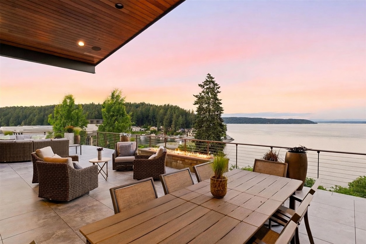 This expansive outdoor deck offers a sophisticated space for entertaining, featuring a dining area with a long table and a comfortable lounge section with wicker furniture. A linear fire feature is integrated into a stone-clad wall, providing a warm focal point against the backdrop of a serene bay and dense forest. The scene is captured at sunset, with the warm, soft light reflecting off the water and creating an inviting, tranquil atmosphere.