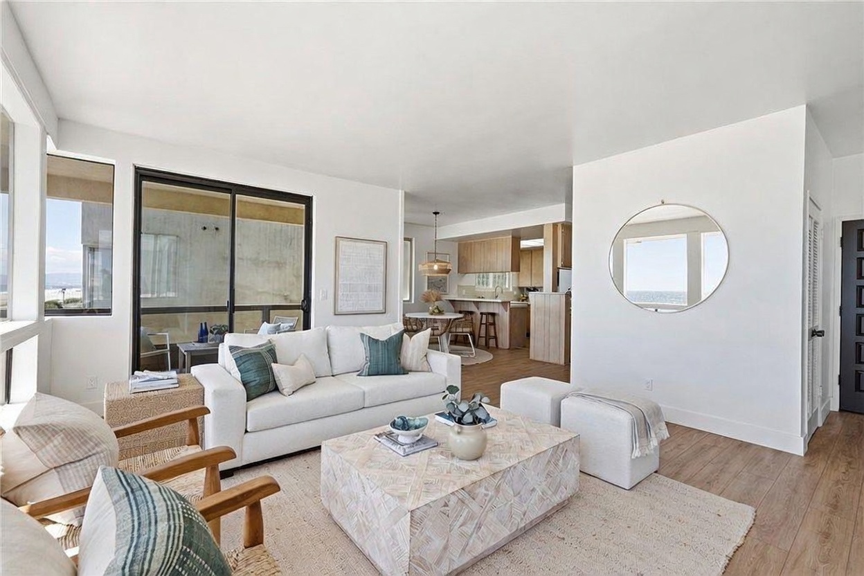 This bright and airy living room features a neutral color palette with light wood-toned flooring and a textured area rug. The space is furnished with a white sofa, two wooden armchairs, and a large, light-toned coffee table, creating a relaxed, coastal-inspired atmosphere. A sliding glass door provides a view of the balcony and ocean, while a circular mirror on the wall reflects the natural light, enhancing the open-concept feel that flows into the adjacent kitchen and dining area.