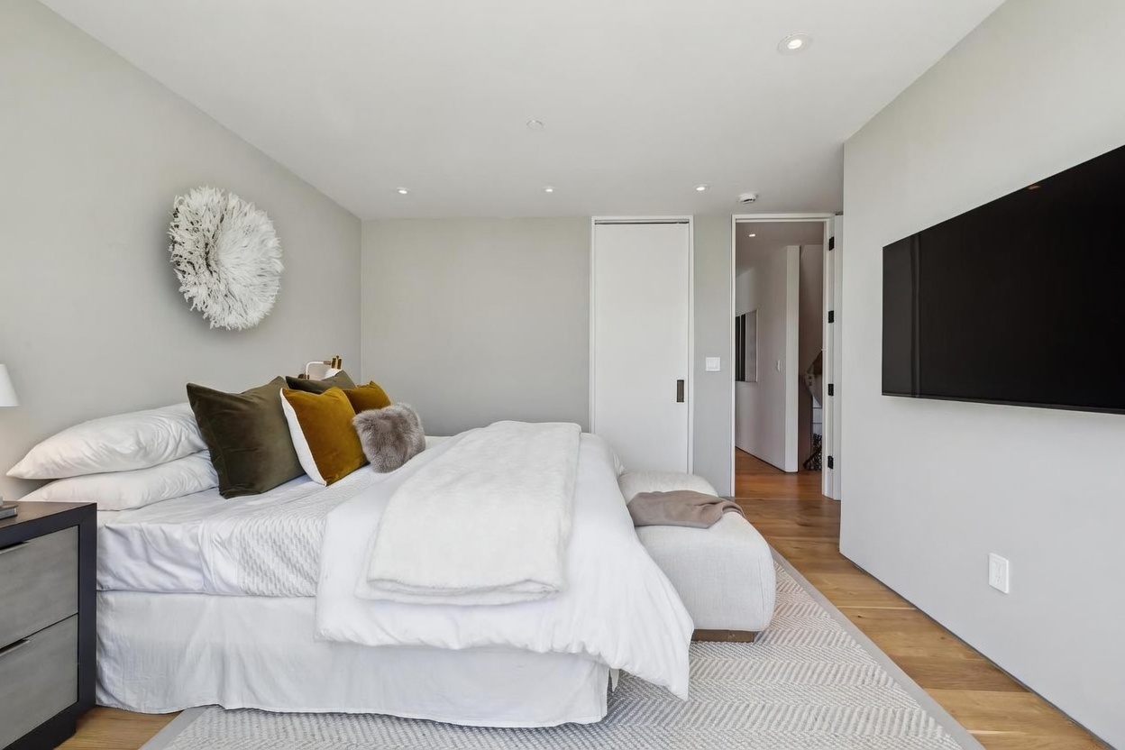 This primary bedroom features a neutral color palette with light gray walls, warm wood flooring, and a textured area rug. The room is anchored by a large bed adorned with white linens and a mix of olive and mustard-toned accent pillows, complemented by a decorative wall hanging above the headboard. A wall-mounted television and a cushioned bench at the foot of the bed complete the space, which opens into a bright hallway.