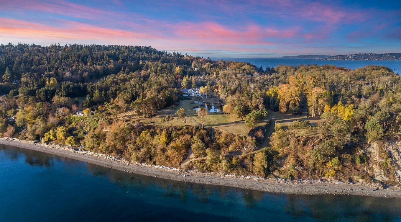 This expansive aerial view captures a secluded coastal property set atop a high bluff overlooking a serene beach and the water. The estate features a large, grassy lawn with a central pond, surrounded by dense, mature forest and autumn-toned trees. The perspective highlights the dramatic topography and the peaceful, private nature of the landscape under a vibrant sunset sky.