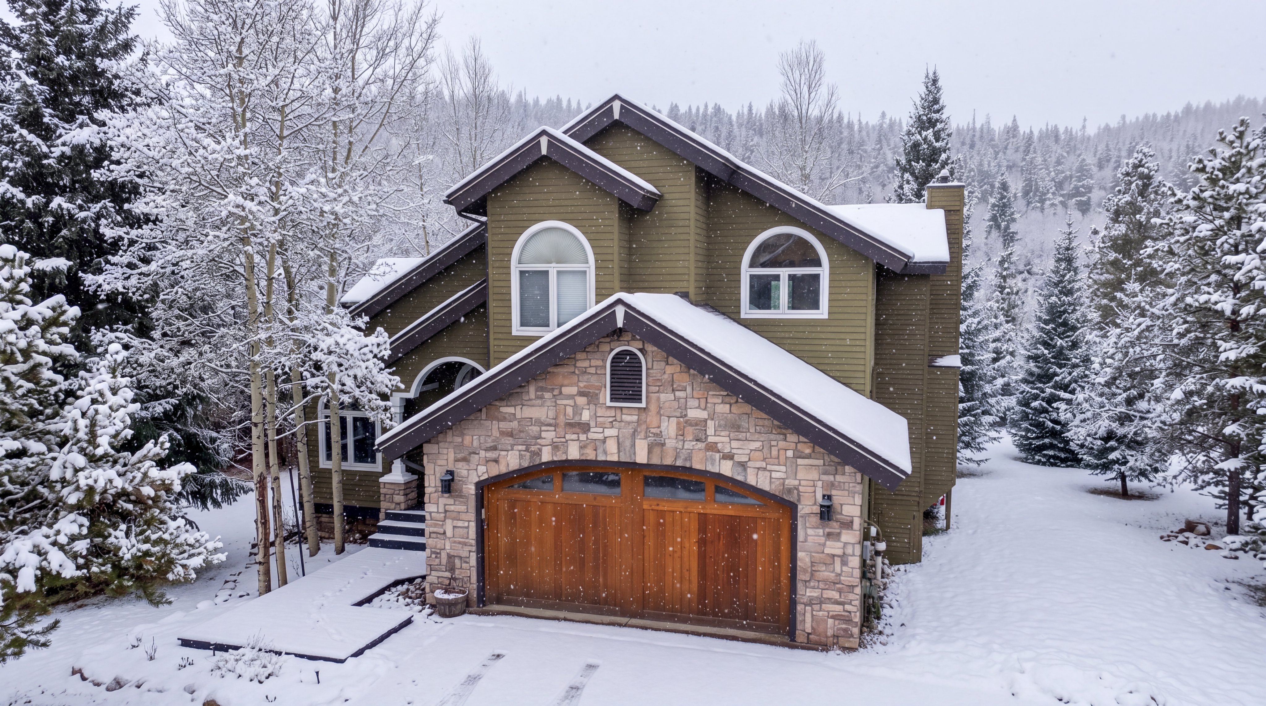 This charming, multi-story home features a striking blend of olive green horizontal siding and natural stone veneer, perfectly suited for a winter setting. The property includes an attached wooden garage with a rustic arched design, surrounded by a picturesque, snow-covered landscape of pine and aspen trees. The elevated aerial angle highlights the architectural rooflines and provides a cozy, serene impression of this mountain-style residence.