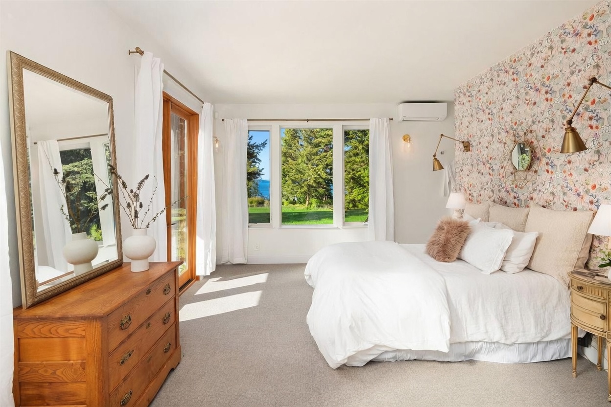 This bright and airy primary bedroom features a striking floral wallpaper accent wall behind the bed, complemented by soft white bedding and neutral carpeting. Large windows provide a scenic view of the lush green trees and water beyond, while a wooden dresser with a large mirror adds a touch of warmth to the space. The room is illuminated by elegant wall-mounted light fixtures, creating a serene and inviting atmosphere.