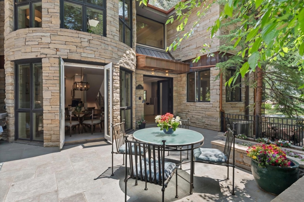 This inviting stone-paved patio features a circular glass-topped dining table surrounded by four metal chairs, creating a perfect outdoor dining space. The area is framed by a distinctive curved stone wall and large glass doors that open into the home's interior, blending indoor and outdoor living. Lush greenery, a vibrant potted plant, and a black metal railing complete the serene, private atmosphere of this outdoor retreat.