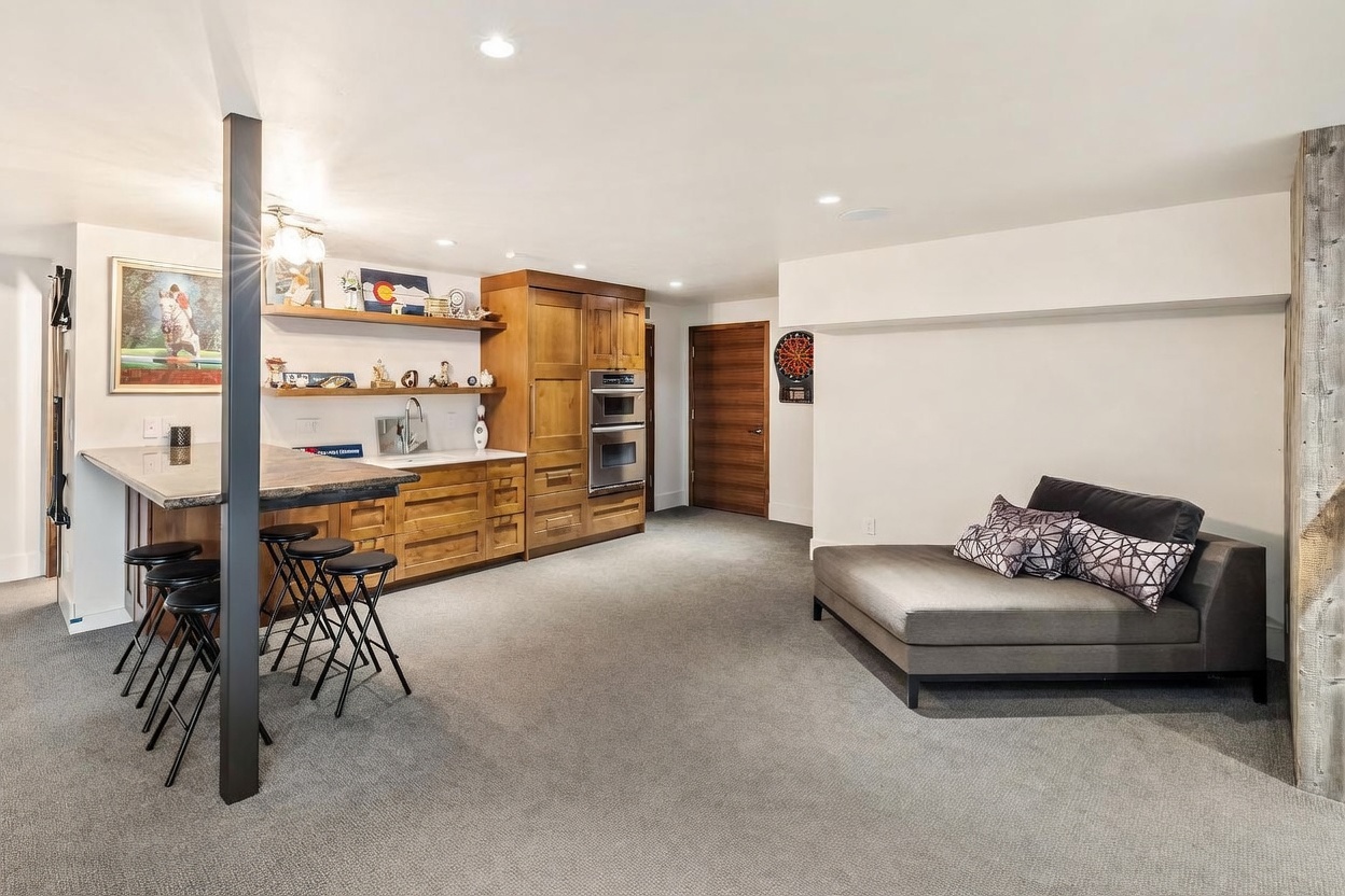 This finished basement features a functional wet bar area with light wood cabinetry, open shelving, and a stone-topped counter with bar seating. The space includes a comfortable lounge area with a grey daybed and a dartboard mounted on the wall, creating a casual entertainment environment. The room is finished with neutral carpeting and recessed lighting, offering a versatile layout for relaxation or hosting guests.