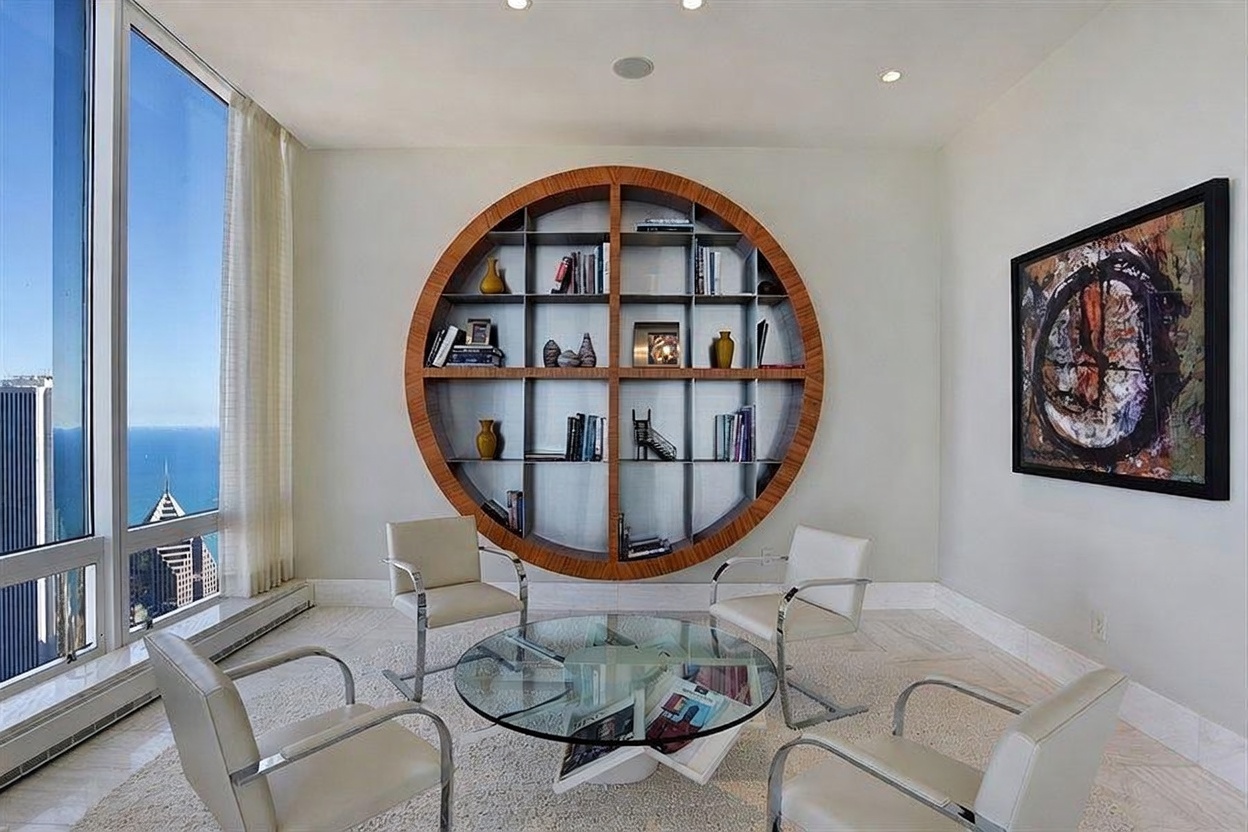 This sophisticated sitting area features a striking circular wooden bookshelf mounted on the wall as a central focal point. Four modern, light-colored armchairs are arranged around a circular glass-topped coffee table, creating an intimate conversation space. Large windows provide an expansive view of the city skyline and water, filling the room with natural light.