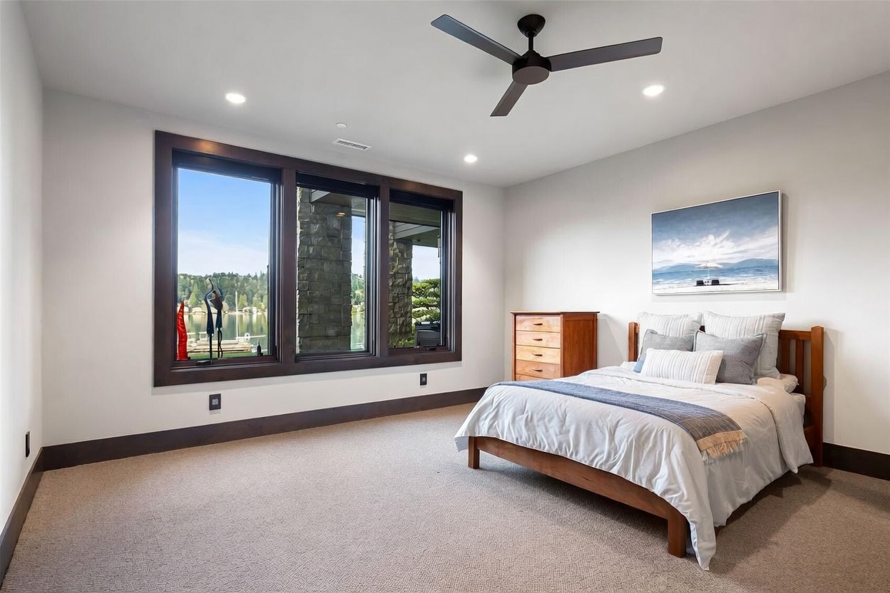 This bright and airy bedroom features neutral walls, light-toned carpeting, and a wooden bed frame dressed in crisp white linens. A large, dark-framed window assembly provides a scenic view of the lake and surrounding greenery, while a wooden dresser sits in the corner. The room is accented by a modern ceiling fan and a piece of coastal-themed wall art, creating a serene and inviting atmosphere.