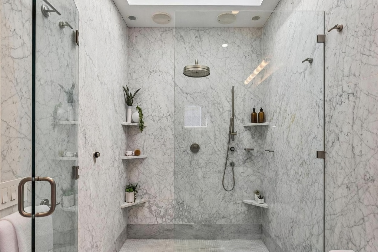 This luxurious walk-in shower features expansive stone-tiled walls with a distinct veined pattern and a matching mosaic-tiled floor. The space is equipped with a rainfall showerhead, a handheld sprayer, and multiple built-in corner shelves for storage, all enclosed by a frameless glass door. The perspective is centered, highlighting the clean, spa-like aesthetic and the high-end fixtures.