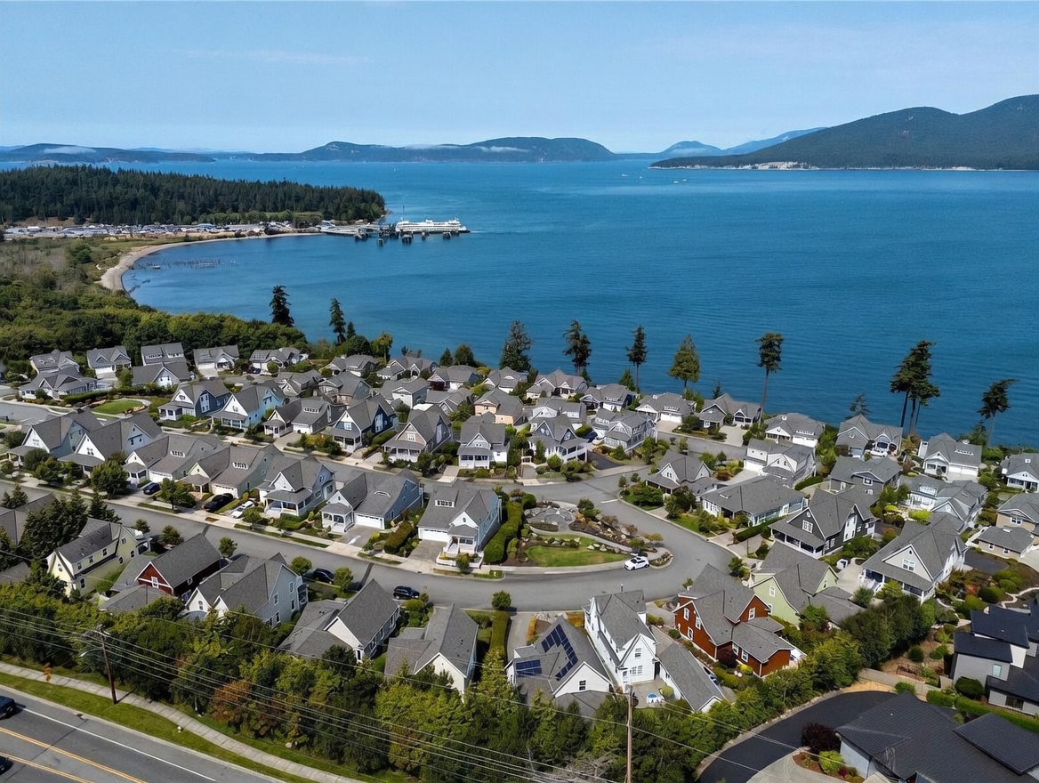 This high-angle aerial view captures a coastal residential neighborhood nestled along a scenic bay. The scene features numerous single-family homes with varied rooflines and neutral siding, organized around winding streets and a central landscaped cul-de-sac. In the background, a ferry terminal extends into the deep blue water, framed by distant mountains and a lush, forested shoreline.