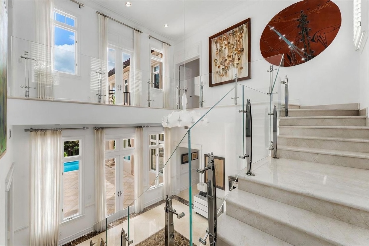 This grand foyer features a sweeping staircase with a glass railing and stone steps, creating an open and airy atmosphere. The double-height space is illuminated by large windows and a unique, sculptural chandelier, while a large circular piece of wall art adds a bold focal point. The perspective from the top of the stairs looks down into the lower level, highlighting the architectural scale and the connection between the two floors.