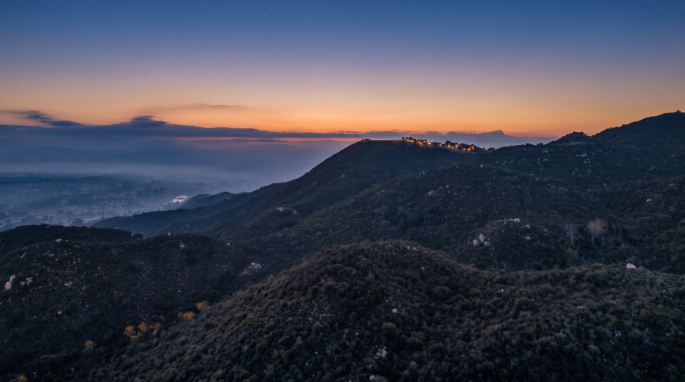 This stunning aerial shot captures a scenic landscape featuring undulating hills covered in dense, natural vegetation during a golden sunrise. In the distance, a misty valley reveals hints of a residential community, creating a serene and picturesque backdrop. The expansive view highlights the property's peaceful hillside location, suggesting privacy and proximity to natural surroundings.