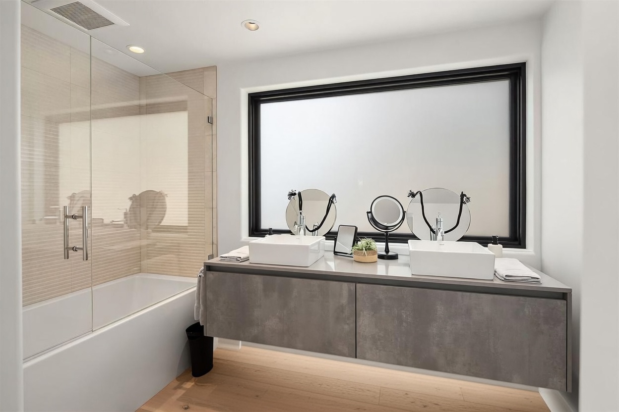 This modern bathroom features a double vanity with square vessel sinks set atop a sleek, grey concrete-textured cabinet. A large, frosted window with a black frame serves as the focal point, flanked by two circular vanity mirrors. To the left, a glass-enclosed shower and tub combination provides a clean, minimalist aesthetic, complemented by light wood-toned flooring.