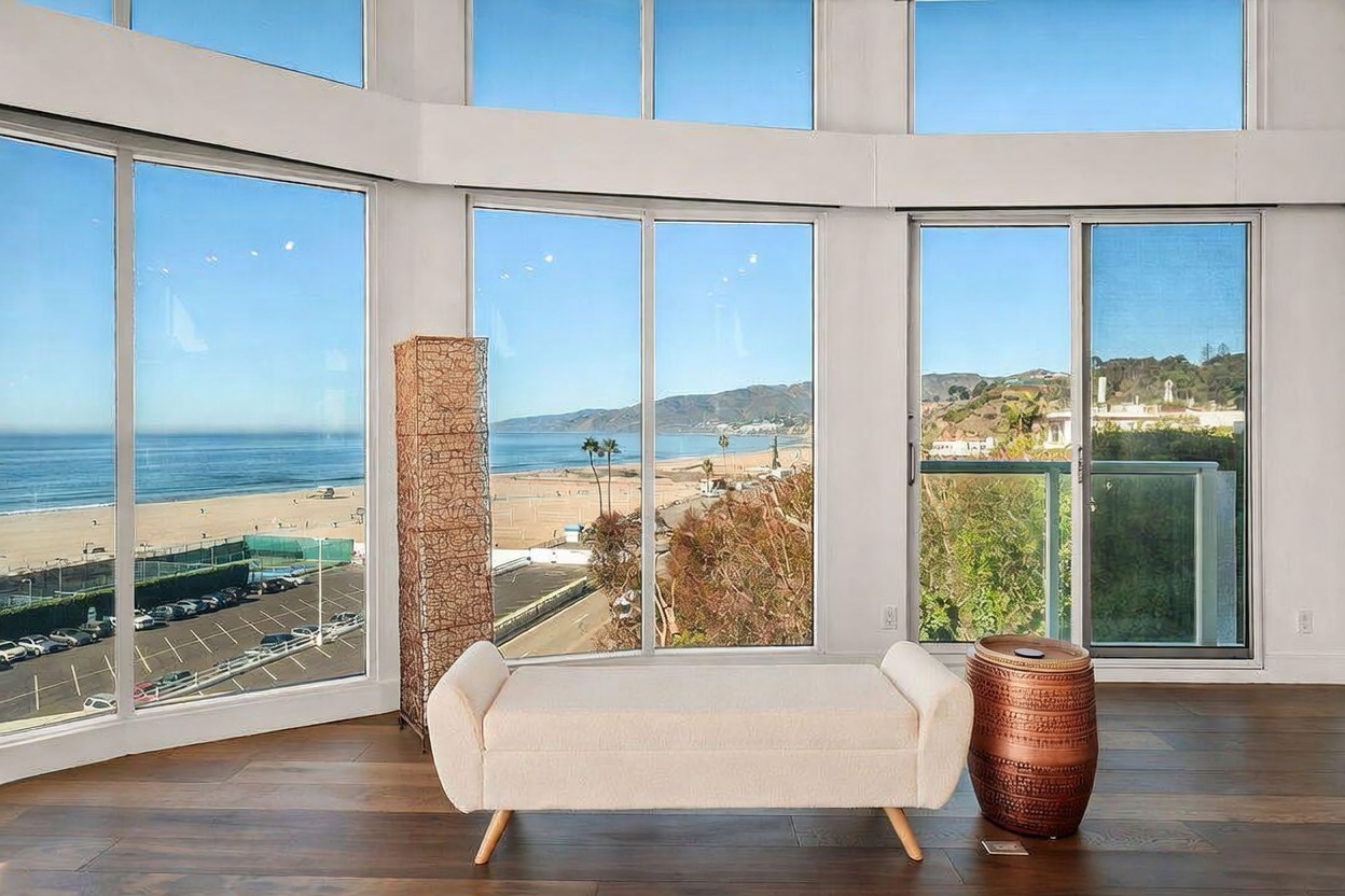 This bright living space features expansive floor-to-ceiling windows that offer a panoramic view of the beach and coastline. The room is furnished with a minimalist light-colored bench and a textured metallic side table, set against a backdrop of warm-toned wood flooring. A tall, woven floor lamp adds a natural element to the airy, sun-drenched interior.