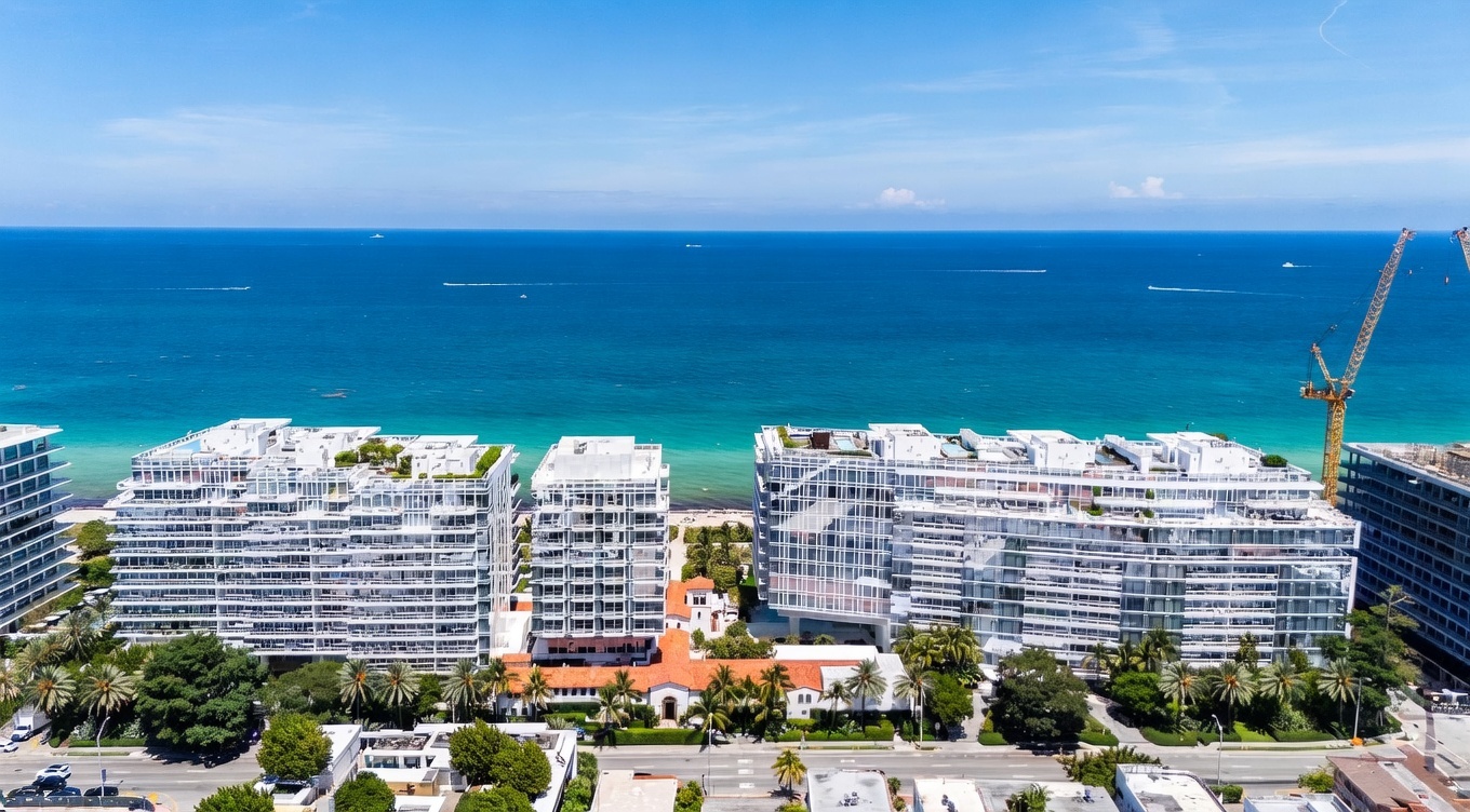 This high-angle aerial view captures a luxury beachfront residential complex featuring modern, multi-story buildings with expansive glass balconies. The property is situated directly along a vibrant turquoise ocean, with lush palm trees and manicured landscaping nestled between the structures. A construction crane is visible on the right, indicating ongoing development in this prime coastal location.