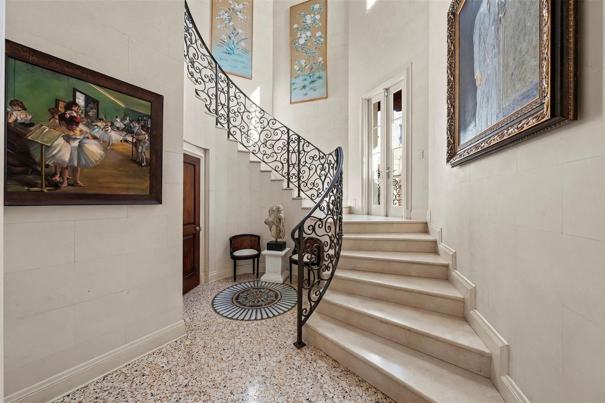 This elegant foyer features a grand curved staircase with an intricate wrought-iron railing that serves as the focal point of the space. The area is adorned with classic artwork, including a large framed painting of dancers and two vertical botanical prints, set against neutral stone-tiled walls. A circular mosaic inlay is set into the speckled stone flooring, creating a sophisticated transition between the entryway and the stairs.