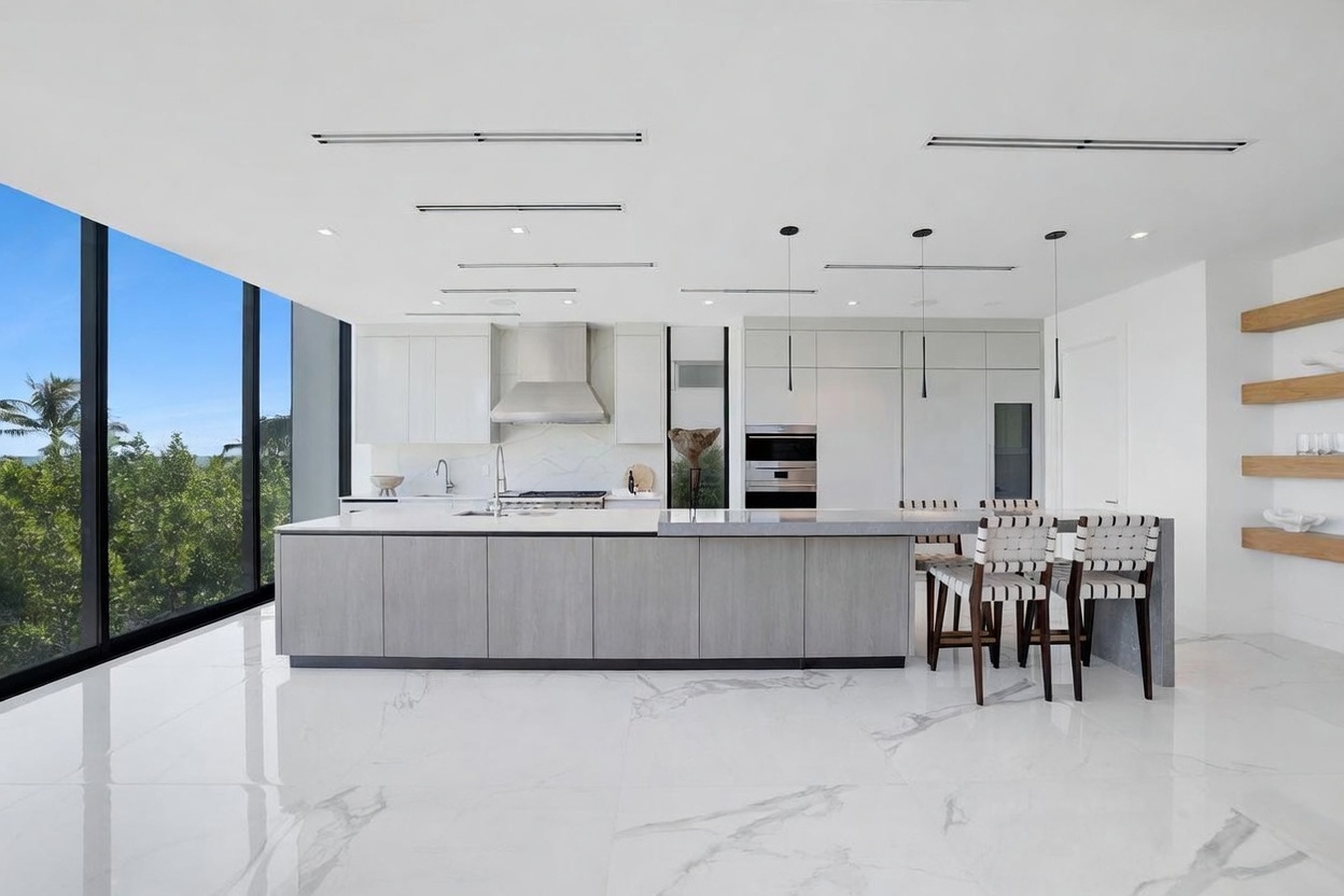 This modern, minimalist kitchen features a large central island with a light-toned wood finish and a sleek, light-colored countertop. The space is defined by clean lines, white cabinetry, and high-end built-in appliances, complemented by a polished, light-colored stone floor. Large windows provide an expansive view of the lush greenery outside, creating a bright and airy atmosphere.