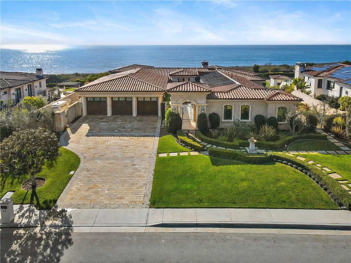 This high-angle, wide-shot perspective captures the front facade of a luxurious Mediterranean-style coastal estate. The property features a three-car garage, a paved stone driveway, and a manicured lawn with structured landscaping, all set against a stunning backdrop of the ocean. The architecture is defined by its warm-toned stucco walls, tiled roof, and an inviting arched entryway.