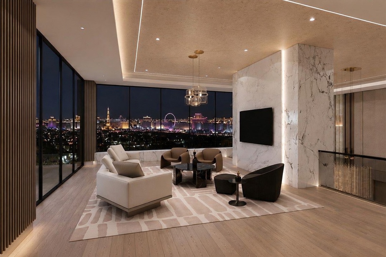 This sophisticated living room features expansive floor-to-ceiling windows that frame a panoramic night view of the city skyline. The space is anchored by a large, light-toned sectional sofa and a pair of curved armchairs arranged on a patterned area rug, centered around a dark stone coffee table. A prominent, full-height stone-clad wall serves as a focal point, while recessed lighting and a modern chandelier provide elegant illumination throughout the room.