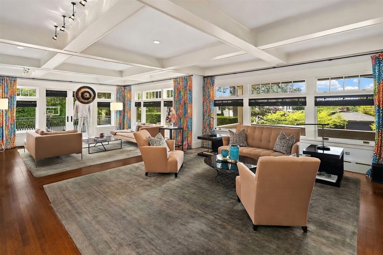 This expansive living room features a coffered ceiling and rich wood flooring, creating a sophisticated and airy atmosphere. The space is furnished with two distinct seating areas, each anchored by large area rugs, and is bathed in natural light from numerous windows adorned with vibrant, multi-colored patterned curtains. The room offers a seamless blend of traditional architectural details and contemporary furniture, providing a spacious and inviting environment for entertaining.