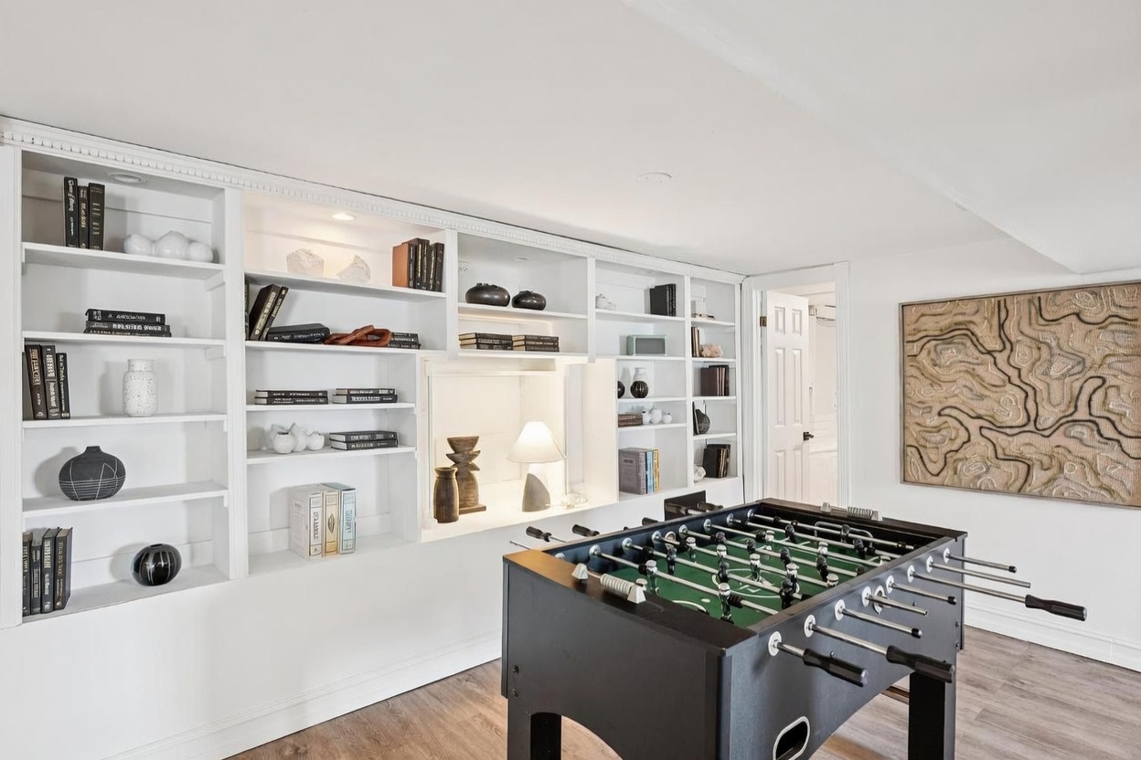 This bright, contemporary basement recreation area features a large, built-in white shelving unit that spans the wall, providing ample space for books and decorative objects. A foosball table sits prominently in the foreground, while a textured, abstract wall art piece hangs on the adjacent wall. The space is finished with light-toned wood-look flooring and clean white walls, creating an inviting and functional entertainment zone.