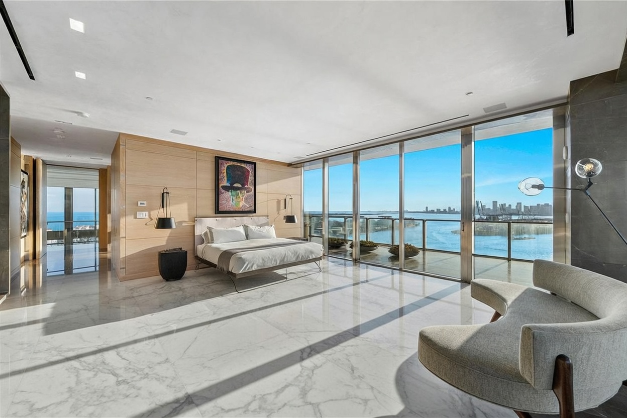 This expansive primary bedroom features a sophisticated, modern design with light wood wall paneling and dark stone flooring. The room is anchored by a large bed facing floor-to-ceiling windows that provide panoramic water and city skyline views, while a seating area with a curved sofa and designer chairs creates a luxurious lounge space. The overall aesthetic is one of high-end, contemporary elegance, enhanced by soft natural light and curated art pieces.