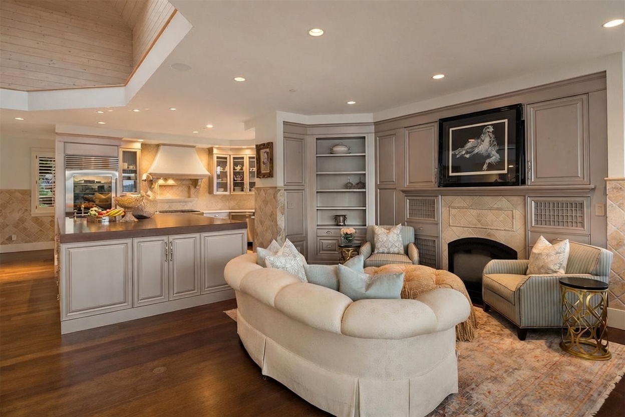 This open-concept living space seamlessly integrates a cozy seating area with a kitchen, featuring warm wood flooring and neutral-toned cabinetry. The living area is anchored by a fireplace with a decorative surround and a framed artwork above, flanked by built-in shelving and two patterned armchairs. A curved sofa faces the fireplace, creating an inviting atmosphere, while the adjacent kitchen island provides a functional transition between the two zones.