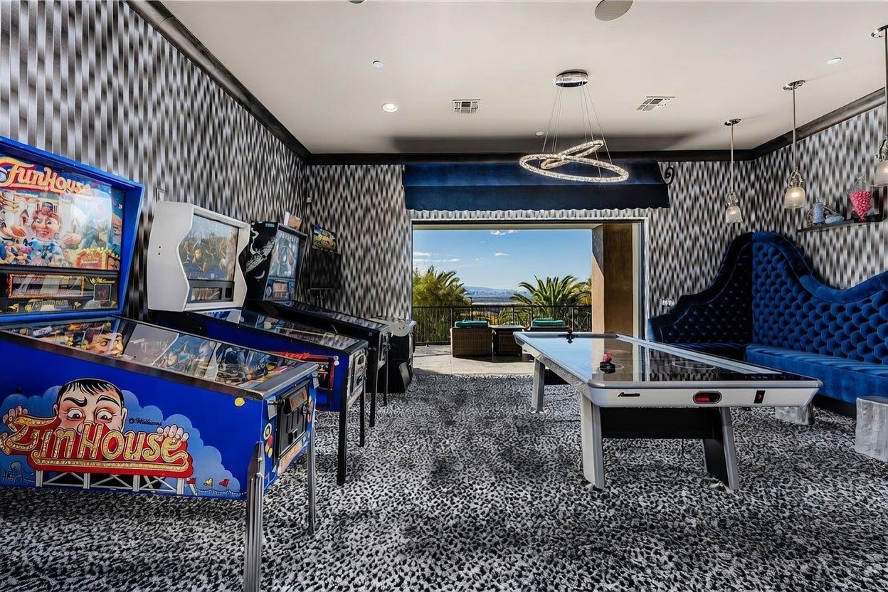 This vibrant game room features a bold, patterned carpet and striking geometric wallpaper, creating a high-energy entertainment space. The room is equipped with two vintage-style pinball machines and a central air hockey table, complemented by a plush, tufted blue velvet sofa along the wall. A large opening leads to an outdoor balcony, providing a scenic view of the surrounding landscape and distant city skyline.