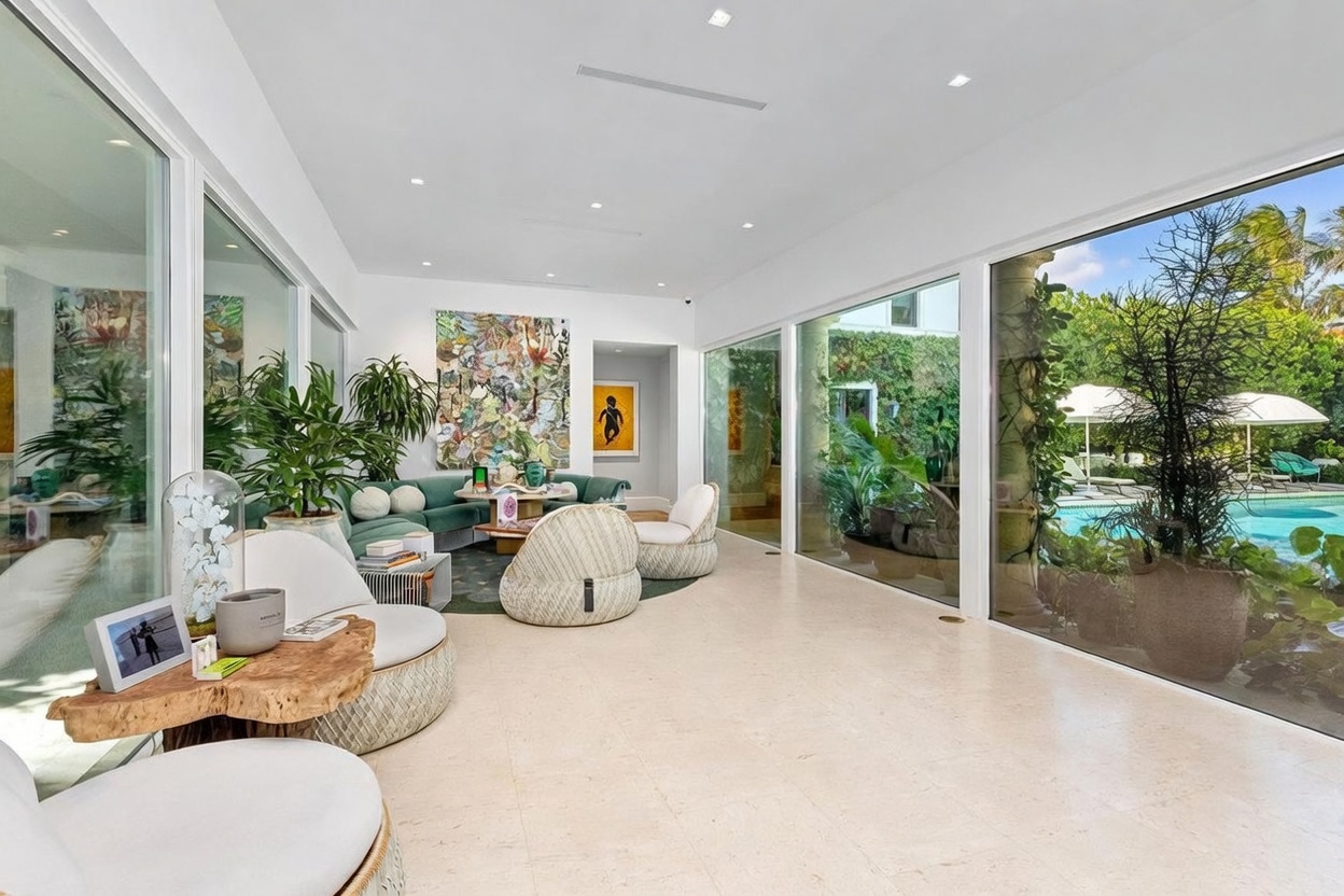 This bright and airy living room features a contemporary design with a neutral color palette and large floor-to-ceiling windows that provide an expansive view of the outdoor pool and lush landscaping. The space is furnished with a curved teal sofa, textured accent chairs, and a unique natural wood-edge coffee table, all arranged on a dark patterned rug. The room is accented by vibrant abstract artwork and indoor greenery, creating a sophisticated and inviting atmosphere.