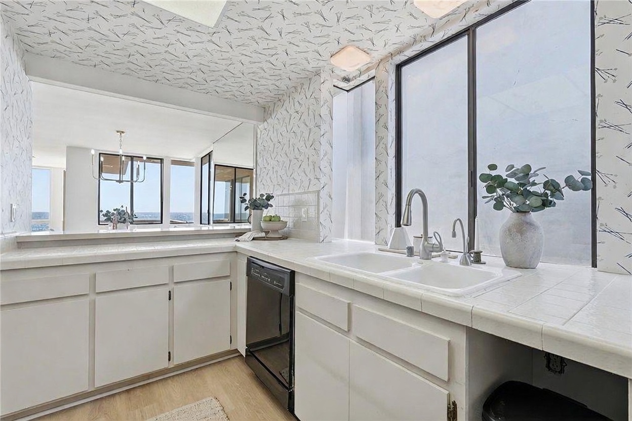 This kitchen features white cabinetry paired with light-colored tiled countertops and a distinctive patterned wallpaper that extends onto the ceiling. A double-basin sink sits beneath a large window that provides a bright, airy feel, while a black dishwasher adds a contrasting element. The space opens up to an adjacent living area with a view of the ocean, creating a seamless connection between the kitchen and the home's coastal setting.