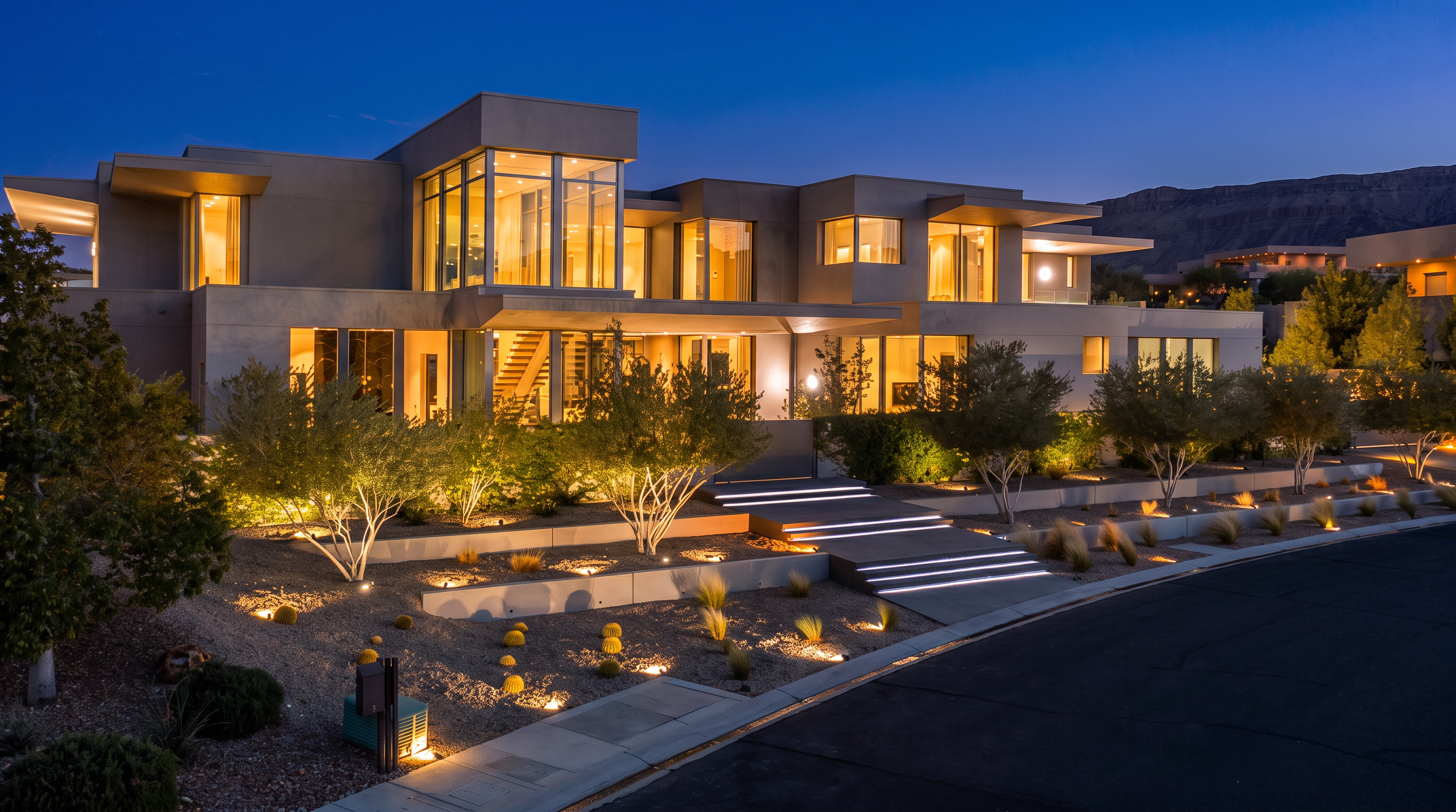 This striking contemporary residence features a multi-level architectural design with clean lines and extensive glass walls that create a seamless indoor-outdoor aesthetic. The front exterior is illuminated by warm landscape lighting, highlighting the terraced desert-style garden, stone retaining walls, and a wide, illuminated staircase leading to the main entrance. Set against a backdrop of distant mountains at dusk, the home exudes a sophisticated, modern luxury appeal.
