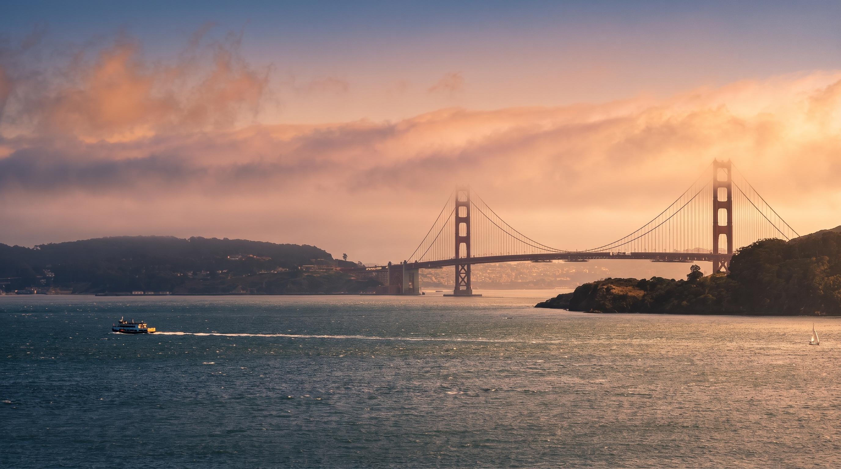 This scenic landscape shot captures the iconic Golden Gate Bridge partially shrouded in low-hanging fog. In the foreground, a ferry boat traverses the choppy blue waters of the bay, while a small sailboat is visible near the rocky shoreline on the right. The perspective is a wide, elevated view that emphasizes the vastness of the bay and the landmark bridge as a prominent feature of the local area.