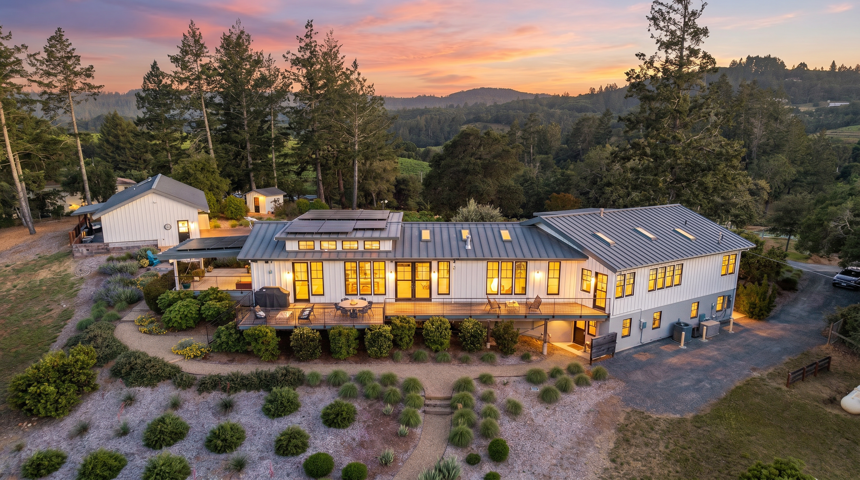 This high-angle aerial perspective captures a modern farmhouse-style residence nestled in a lush, hilly landscape. The home features a metal roof with integrated solar panels, white siding, and a spacious wooden deck overlooking a meticulously landscaped garden with gravel pathways and manicured shrubs. A separate detached garage and a scenic backdrop of dense forest and rolling hills complete this serene, rural property.