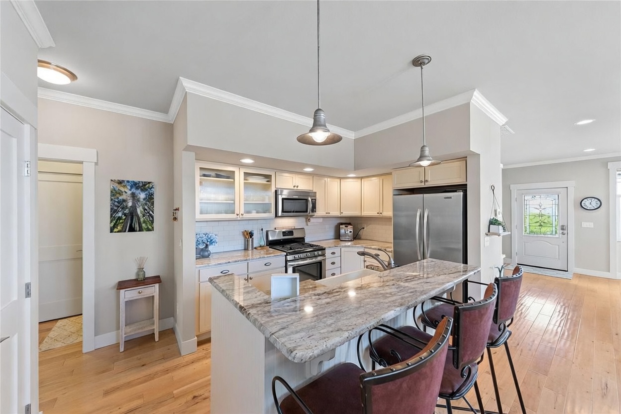 This bright, open-concept kitchen features light-toned cabinetry, a stone-topped island with bar seating, and stainless steel appliances. The space is illuminated by dual pendant lights hanging over the island and recessed ceiling lighting, creating a warm and inviting atmosphere. The kitchen flows seamlessly into the adjacent living area, showcasing light wood flooring and a neutral color palette throughout.