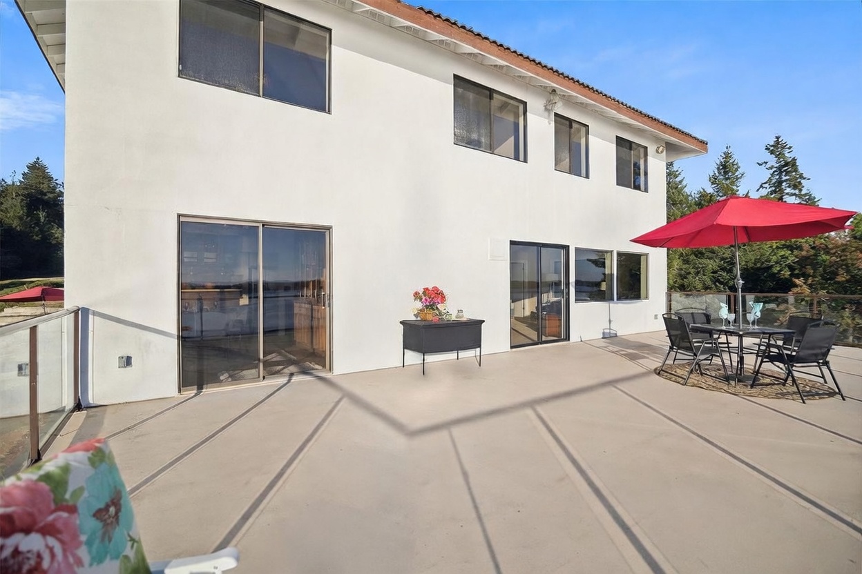 This spacious, elevated deck features a smooth, light-colored surface and a modern glass railing that provides an unobstructed view of the water beyond. A dining set with a vibrant red umbrella sits to the right, while a small console table with a floral arrangement adds a touch of decor near the sliding glass doors. The white exterior wall of the home is punctuated by several large windows, creating a bright and airy atmosphere perfect for outdoor entertaining.