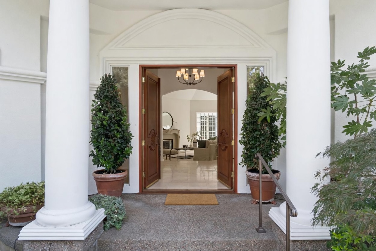 This grand entryway features stately white columns and a striking set of wooden double doors that open into a well-lit home interior. The scene is framed by manicured topiaries in classic terracotta pots and a welcoming front mat, creating an inviting and elegant first impression. The architectural details, including the arched transom window above the entrance, convey a sense of traditional refinement.
