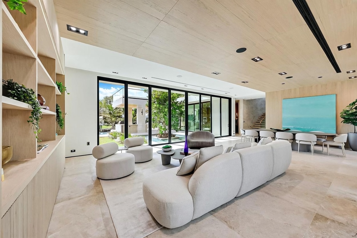 This expansive, modern living room features a neutral color palette with light wood ceiling paneling and large, floor-to-ceiling glass sliding doors that open to a lush outdoor courtyard. The space is furnished with a curved, light-toned sectional sofa, two matching lounge chairs, and a minimalist coffee table, all set upon a soft area rug. A long dining table is visible in the background, and a large, vibrant blue abstract painting serves as a focal point on the wall, creating a bright and airy atmosphere.