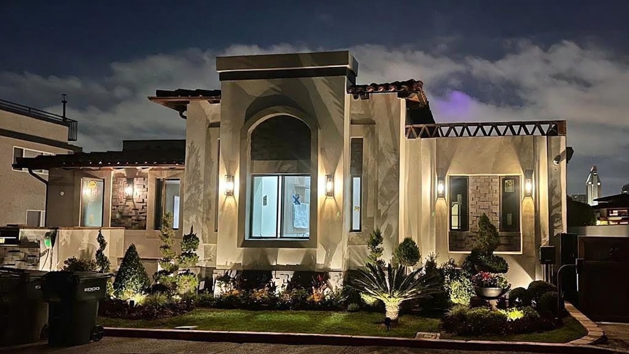 This evening shot captures the front facade of a contemporary home with a stucco exterior and warm, integrated lighting. The architecture features a prominent central arched window, stone-accented wall sections, and a decorative geometric frieze along the roofline. The landscaped front yard includes manicured shrubs and a small palm tree, creating an inviting and sophisticated curb appeal.