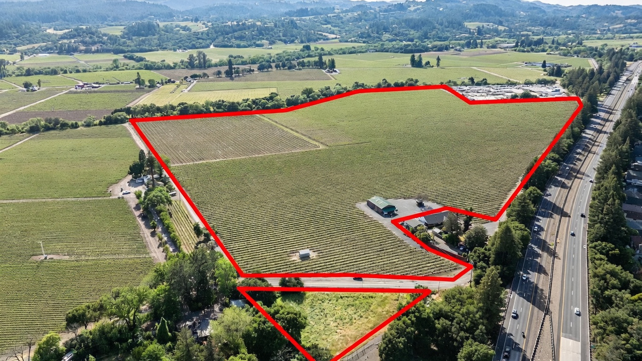 This high-angle aerial view showcases a sprawling vineyard property outlined in red, highlighting its expansive agricultural land and proximity to a major highway. The site includes a cluster of buildings, including a barn-style structure and a main residence, set amidst rows of grapevines. The perspective emphasizes the vast scale of the estate and its convenient access to regional transportation routes.