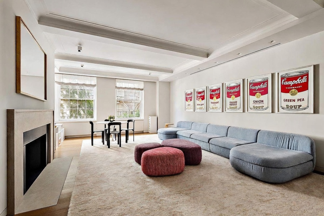 This spacious living room features a large, light-blue modular sofa arranged against a wall adorned with a series of six framed Campbell's soup can prints. A pair of textured, reddish-maroon ottomans sits on a plush, neutral-toned area rug, while a small dining table and chairs are positioned near the tall windows. The room is anchored by a minimalist fireplace with a tall, light-colored surround, creating a sophisticated and art-focused atmosphere.