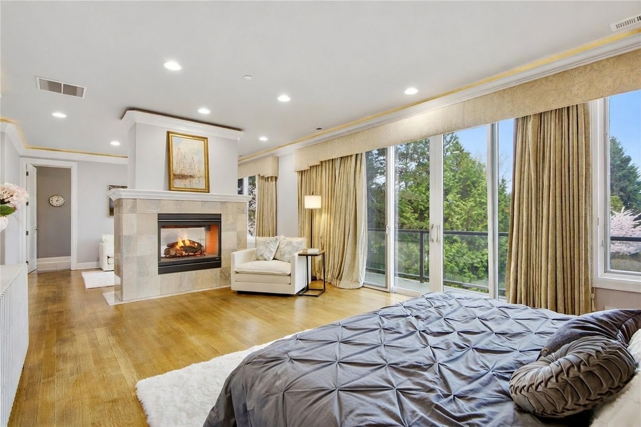 This spacious primary bedroom features light wood flooring and a central stone-clad chimney breast that separates the sleeping area from a cozy sitting nook. Large glass sliding doors provide access to a balcony and offer expansive views of the surrounding greenery, while soft, neutral-toned curtains frame the windows. The room is elegantly appointed with a plush bed, a comfortable armchair, and recessed lighting, creating a serene and luxurious atmosphere.