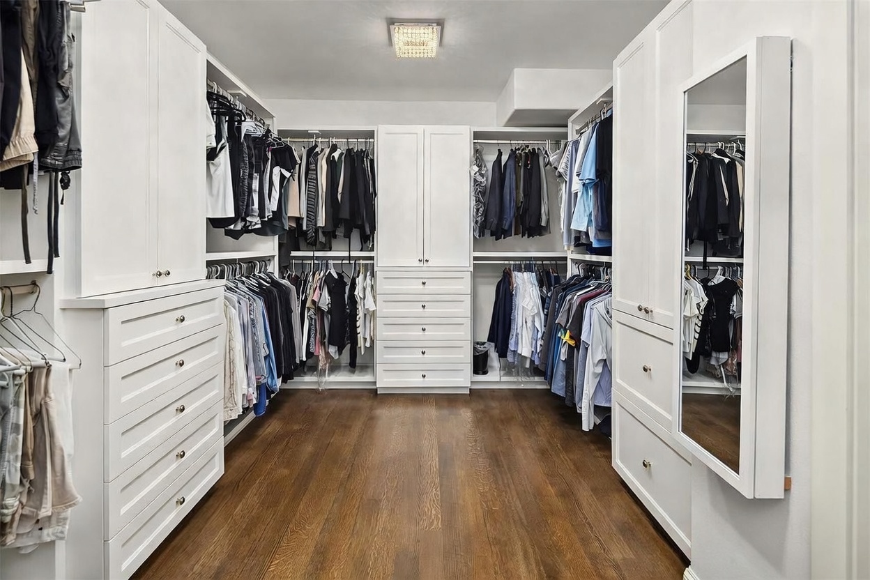This spacious walk-in closet features custom white cabinetry with extensive hanging space, open shelving, and multiple drawers for organized storage. The room is anchored by dark wood flooring and illuminated by a central crystal-style light fixture, creating a bright and polished aesthetic. A full-length mirror is integrated into the cabinetry on the right, enhancing the functionality and depth of the space.