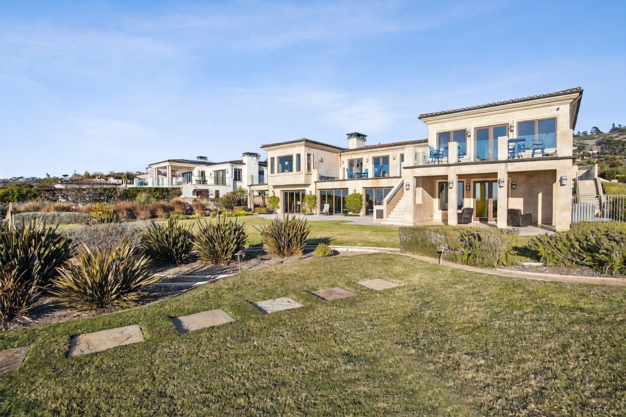This image captures the expansive rear exterior of a large, multi-story Mediterranean-style estate. The home features a light-colored stucco facade, multiple balconies with glass railings, and a series of large windows and glass doors that open onto a stone patio. A well-manicured lawn with stone pavers leads up to the residence, set against a clear blue sky and a distant hillside landscape.