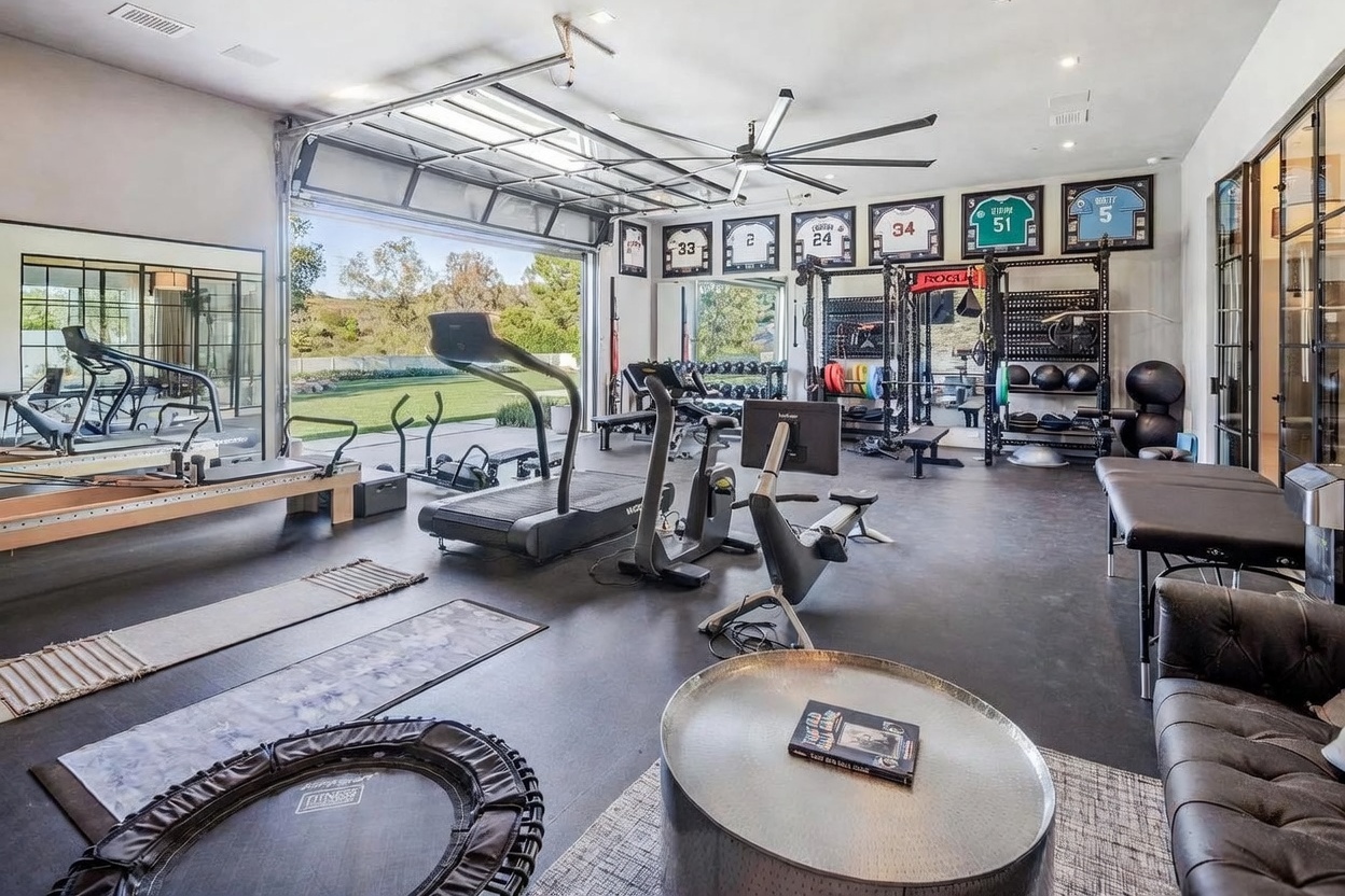 This expansive home gym features a modern, industrial aesthetic with a large, open-air garage door that seamlessly connects the workout space to the outdoor landscape. The room is equipped with professional-grade fitness gear, including a treadmill, rowing machine, Pilates reformer, and a comprehensive Rogue weight rack adorned with framed sports jerseys on the wall. A sleek, circular metal coffee table sits in the foreground, adding a touch of lounge-style comfort to this high-end athletic facility.