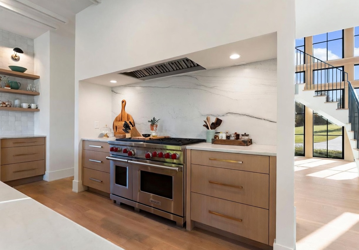 This modern kitchen features light wood cabinetry with sleek hardware and a prominent stainless steel range with red knobs. A large stone backsplash provides a clean, elegant backdrop, while open shelving to the left adds a touch of warmth. The perspective looks directly at the cooking area, with a glimpse of a staircase and large windows visible in the adjacent space.