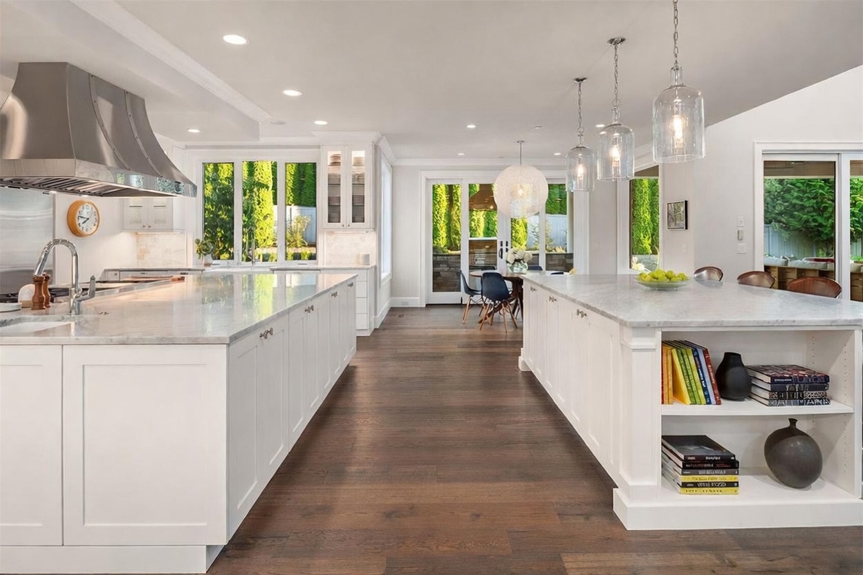 This bright and spacious kitchen features dual islands with stone countertops and white cabinetry, creating a symmetrical and functional layout. The room is anchored by dark wood flooring and illuminated by a combination of recessed lighting and elegant glass pendant lights. Large glass doors at the rear provide a seamless transition to the outdoor dining area, enhancing the open-concept feel of the home.